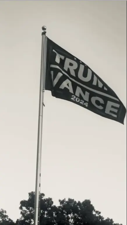 A black Trump Vance 2024 flag waving on a flagpole against a clear sky, with bold white text showing support for the 2024 campaign.